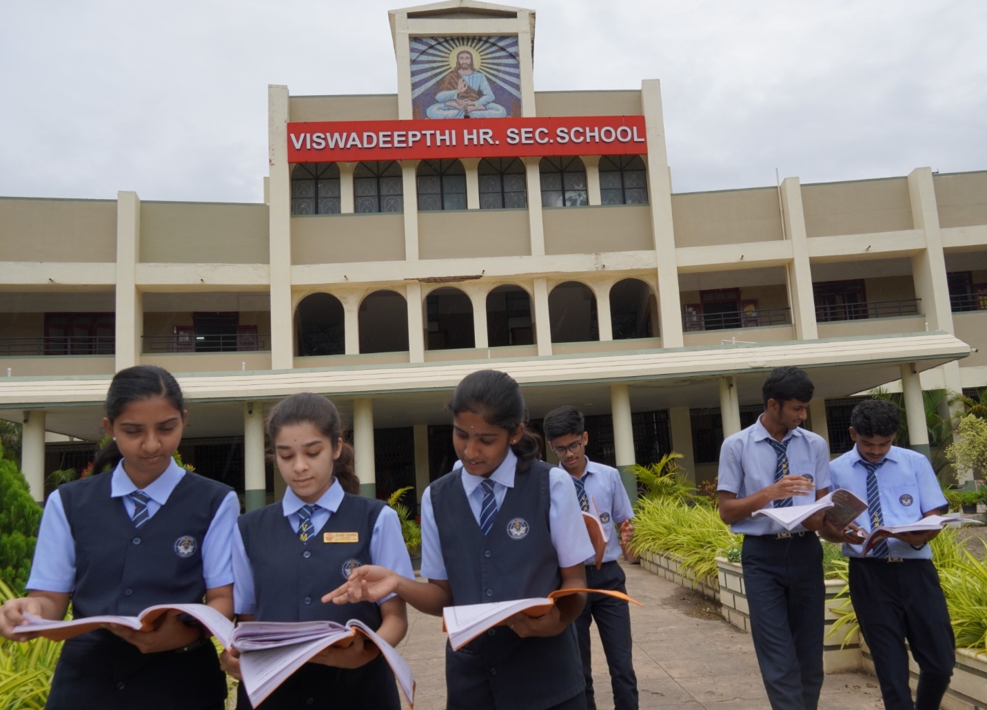 Group of students in school uniforms standing outside Viswadeepthi Hr Sec School, reading books together in front of a large building with a red sign that reads VISWADEEPTHI HR SEC SCHOOL and a painting above the entrance, surrounded by greenery, conveying a focused and collaborative atmosphere
